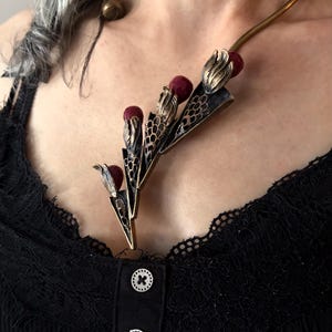 May include: A gold-toned metal collar necklace featuring three stylised floral elements with dark red spherical beads and intricate openwork mesh detailing. The necklace rests against a black ribbed top with three white buttons and lace trim.