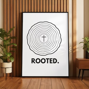 Könnte beinhalten: Ein rechteckiger Wandkunstdruck in einem schwarzen Rahmen mit weißem Hintergrund und einer schwarzen Strichzeichnung von Baumringen. In der Mitte der Ringe befindet sich ein kleines schwarzes Kreuz. Unter der Grafik steht das Wort ROOTED. in schwarzen Großbuchstaben.