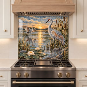 Op de afbeelding: Een keukenachterwand met een mozaïek-stijl illustratie van twee kraanvogels in een moeras bij zonsondergang. De scène bevat witte waterlelies, hoge lisdodden en een gouden zon die reflecteert op het water, geplaatst achter een roestvrijstalen fornuis.
