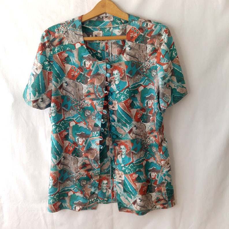 May include: A teal and orange button-up shirt with a short sleeve and a colorful print of faces and abstract shapes. The print includes the words "MRS" and "HAT".