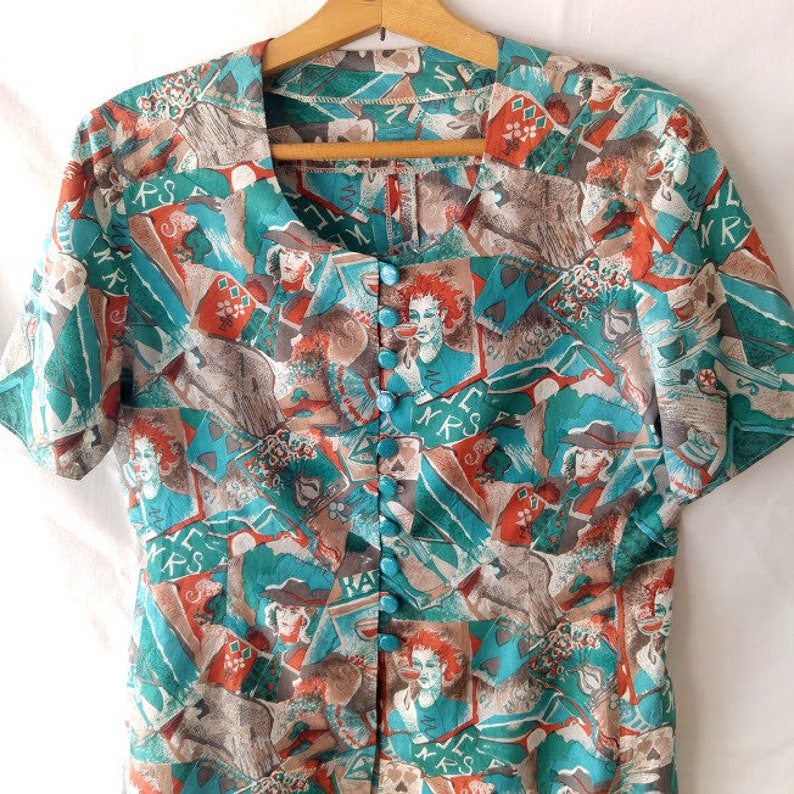 May include: A turquoise and brown short-sleeved button-up blouse with a colorful abstract print. The print features various figures, letters, and symbols. The blouse has a round neckline and a button-up front.