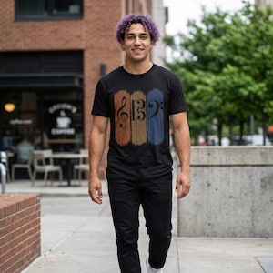Könnte beinhalten: Ein schwarzes Kurzarm-T-Shirt mit einem Grafikdruck aus drei vertikalen, abgenutzten rechteckigen Feldern in Rost, Beige und Blau, die jeweils ein schwarzes Notenschlüssel-Symbol zeigen: einen Violinschlüssel, einen C-Schlüssel und einen Bassschlüssel.