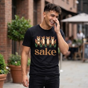 May include: A black short-sleeved t-shirt featuring a graphic of four cartoon foxes standing in a row above green fern leaves. Below the foxes, the word sake is printed in a large, bold, tan-colored sans-serif font across the chest area.