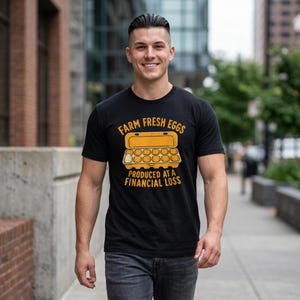 Könnte beinhalten: Ein schwarzes Kurzarm-T-Shirt mit einer gelben Grafik eines offenen Eierkartons, der ein einzelnes Ei enthält. Der Text über dem Karton lautet FARM FRESH EGGS und der Text darunter lautet PRODUCED AT A FINANCIAL LOSS in fetter gelber Schrift.