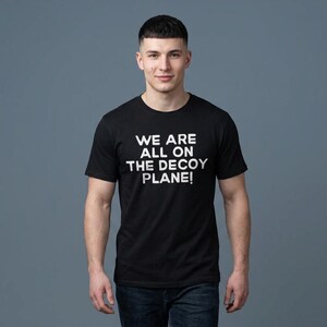May include: A black short-sleeved t-shirt featuring the white text WE ARE ALL ON THE DECOY PLANE! printed in a bold, sans-serif font across the chest. The garment has a classic crew neckline and a standard fit, providing a simple and clean look.