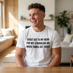 May include: A white short-sleeved t-shirt featuring black text printed on the chest that reads: SHOUT OUT TO MY MOM FOR NOT STRANGLING ME WHEN THINGS GET TOUGH. The garment has a classic crew neckline and a standard fit, made of a plain white fabric.