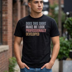 May include: A black short-sleeved t-shirt featuring the text "DOES THIS SHIRT MAKE ME LOOK PROFESSIONALLY DEVELOPED" printed in a stacked layout. The words "DOES THIS SHIRT MAKE ME LOOK" are in grey, "PROFESSIONALLY" is in pink, and "DEVELOPED" is in tan.