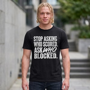 May include: A black short-sleeved t-shirt featuring white distressed-style text that reads: STOP ASKING WHO SCORED. ASK WHO BLOCKED. The lettering is arranged in a block format with the word WHO emphasized in a larger, slanted script font.