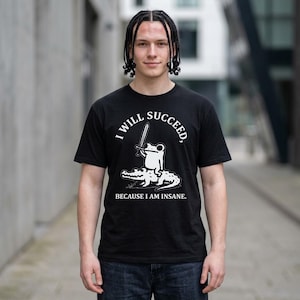 Puede incluir: Una camiseta negra de manga corta con un gráfico blanco de una rana sosteniendo una espada mientras monta un caimán. El texto "I WILL SUCCEED, BECAUSE I AM INSANE." está impreso en letras mayúsculas blancas sobre y bajo la ilustración.