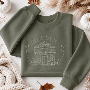Könnte beinhalten: Ein olivgrünes Sweatshirt mit Rundhalsausschnitt und einer weißen Strichzeichnung eines Café-Ladens. Der Text GOOD DAYS steht über dem Laden und START WITH COFFEE darunter, ergänzt durch ein kleines Herzsymbol in der Mitte unter dem Design.