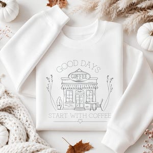 Könnte beinhalten: Ein weißes Sweatshirt mit Rundhalsausschnitt und einer schwarzen Strichzeichnung eines Cafés. Der Text GOOD DAYS steht über dem Café, COFFEE auf dem Schild und START WITH COFFEE darunter, ergänzt durch ein kleines Herzsymbol am unteren Rand.