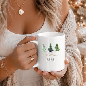 Könnte beinhalten: Ein weißer Keramikbecher mit einer Aquarellillustration von drei grünen Tannenbäumen. Unter den Bäumen steht der Text Merry Christmas in goldener Schreibschrift, gefolgt von dem Wort MAMA in einer einfachen, dunklen, serifenlosen Großschrift.