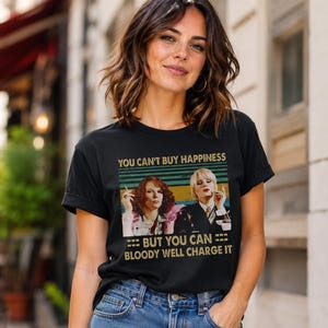 May include: A black short-sleeved t-shirt featuring a graphic print of two women with cigarettes. The text reads: YOU CAN'T BUY HAPPINESS BUT YOU CAN BLOODY WELL CHARGE IT. The design includes horizontal stripes in muted green, yellow, and blue tones.