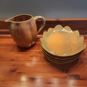 May include: A ceramic pitcher with a glossy, mottled amber and brown finish alongside a stack of four matching leaf-shaped plates. The pitcher features a curved handle and a spout, while the plates have embossed vein details in a warm, earthy tone.