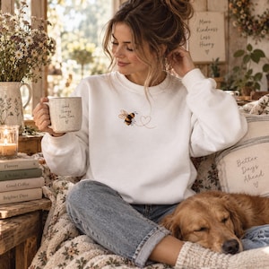 Könnte beinhalten: Ein weißes Sweatshirt mit Rundhalsausschnitt und einer kleinen Stickerei einer Hummel, die auf eine gepunktete Herzform zufliegt. Das Sweatshirt wird mit einer blauen Jeans kombiniert. Eine Keramiktasse mit dem Text Good Things Take Time wird gehalten.