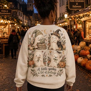 Könnte beinhalten: Ein cremefarbenes Sweatshirt mit einer Grafik auf der Rückseite, die vier Cartoon-Geister umgeben von Kürbissen, Blumen und Sternen zeigt. Der Text lautet, A little spooky, a lot of feelings. Die Geister schlafen, schauen grimmig mit einer Katze, halten Blumen und schwingen einen Zauberstab.