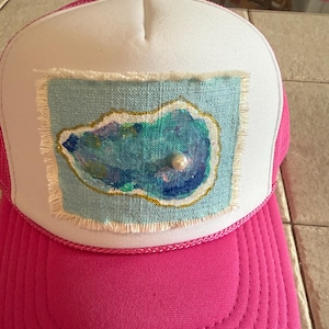 May include: A white and bright pink trucker hat featuring a rectangular fabric patch with frayed edges. The patch displays a painted blue and green oyster shell design with a gold outline and a single white pearl bead attached to the center.