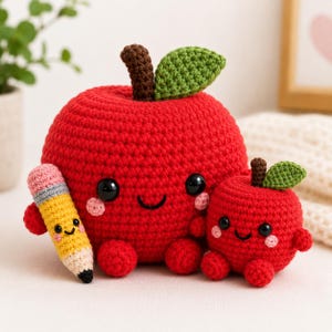 May include: Two crocheted red apples with smiling faces, black bead eyes, and pink cheeks. The larger apple has a brown stem and green leaf, holding a yellow, pink, and grey crocheted pencil. The smaller apple also has a brown stem and green leaf.