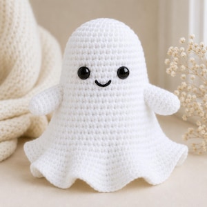 May include: A white crocheted ghost plush toy featuring a rounded head, two small arms, and a flared base. The ghost has two round, glossy black plastic safety eyes and a simple, stitched black curved line for a mouth, creating a friendly expression.