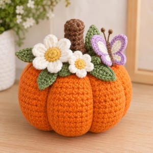 May include: A crocheted orange pumpkin featuring a brown stem, two white flowers with yellow centers, several green leaves, and a purple and white butterfly with brown antennae. The pumpkin is crafted with a textured yarn stitch pattern.