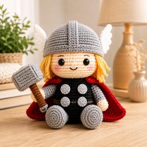 May include: A crocheted doll featuring a character with yellow yarn hair, a grey helmet with white wings, a black torso with grey circular accents, and a red cape. The doll holds a grey crocheted hammer with a brown handle in its right hand.