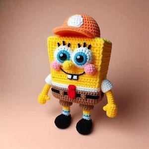 Op de afbeelding: Een handgemaakte gehaakte pop in de vorm van een rechthoekig geel figuur met grote blauwe ogen, lange wimpers en een brede glimlach. Het figuur draagt een oranje pet, een wit overhemd, een bruine broek met een rode das en zwarte schoenen met witte sokken.