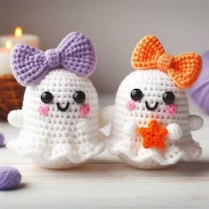 May include: Two white crocheted ghost figures with black bead eyes and pink stitched cheeks. One ghost features a lavender crocheted bow on its head, while the other has an orange crocheted bow and holds a small orange crocheted star on its front.