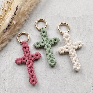 Può includere: Tre portachiavi a forma di croce in macramè nei colori rosa antico, verde salvia e crema. Ogni croce è realizzata in corda annodata e presenta un anello metallico circolare color oro sulla parte superiore per l'aggancio. I design sono uniformi.