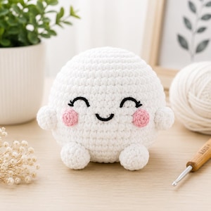 May include: A round, white crocheted plush toy featuring a smiling face with black embroidered eyes and a mouth, along with two small pink circular cheeks. The plush has two small rounded arms and two small rounded feet attached to its body.