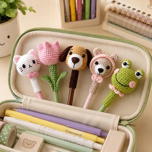 May include: A light green zippered pencil case containing five crochet pen toppers shaped like a white cat, a pink tulip, a brown dog, a beige bear, and a green frog. The case also holds several pens, a small bear-printed eraser, and two rolls of washi tape.