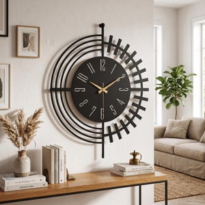 Könnte beinhalten: Eine große, runde Wanduhr mit schwarzem Zifferblatt sowie goldenen Ziffern und Zeigern. Die Uhr ist von einem dekorativen schwarzen Metallrahmen umgeben, der aus konzentrischen Halbkreisen links und vertikalen Stäben rechts besteht.
