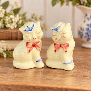 Peut inclure: Une paire de salières et poivrières en céramique en forme de chat de couleur crème. Chaque chat présente un nœud rouge peint à la main autour du cou, un petit papillon bleu sur la tête et des détails bleus sur la patte. Yeux fermés et moustaches.