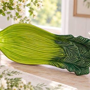 May include: A ceramic serving platter shaped like a head of bok choy, featuring a glossy finish with a gradient of light green at the stem transitioning to a deep, dark forest green at the textured, ruffled leaf edges, creating a realistic vegetable form.