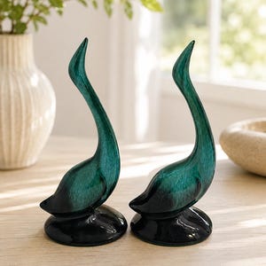 May include: Two matching swan figurines featuring a glossy, deep emerald green finish that transitions into a solid black base. Each bird is depicted with a long, slender, curved neck and a smooth, streamlined body, standing on a circular black pedestal.