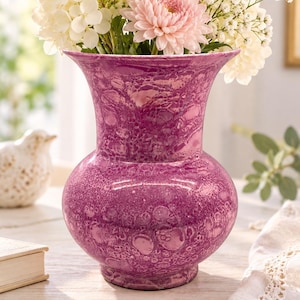 May include: A bulbous ceramic vase with a flared rim and a vibrant magenta glaze featuring a textured, mottled pattern of lighter and darker purple spots. The vase is filled with a bouquet of white hydrangeas and a single pale pink chrysanthemum.