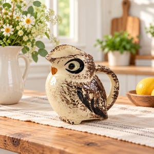 May include: A ceramic jug shaped like an owl with a cream-coloured body and brown speckled details. The owl features large black and white eyes, a small orange beak, and a curved handle on the side. The jug has a wide, open pouring spout.