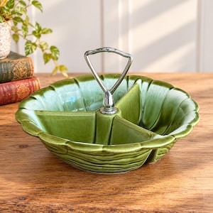 May include: A round, three-sectioned green ceramic serving dish with a scalloped edge and a glossy finish. The dish features a central silver-colored metal handle with a triangular loop for carrying, attached to the middle of the bowl.