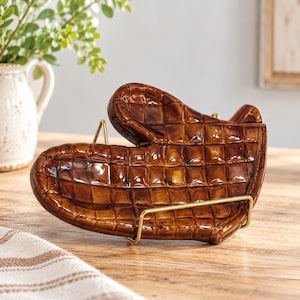 May include: A brown ceramic oven mitt shaped spoon rest with a glossy finish and a quilted grid pattern. The spoon rest is held upright by a thin gold-toned metal wire stand, showcasing its detailed texture and curved shape.