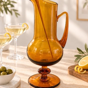 May include: A tall amber-coloured glass pitcher with a rounded body, a slender neck, a curved handle, and a matching glass stirring rod. The pitcher stands on a circular pedestal base and features a wide pouring spout at the top rim.