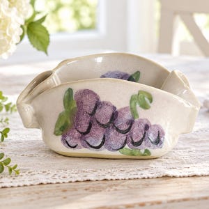 May include: A cream-coloured ceramic bowl with a hand-painted purple grape cluster design on the exterior. The bowl features a wide, open shape with curved, integrated handles on both sides and a smooth, speckled finish throughout the surface.