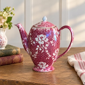 Puede incluir: Una tetera de cerámica con un patrón floral en color magenta intenso y blanco, con un acabado texturizado de líneas entrecruzadas. La tetera incluye una tapa a juego con un pequeño pomo, un asa curva y un pico vertedor cónico.