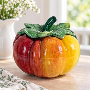 May include: A ceramic pumpkin-shaped container featuring a glossy finish with a gradient of colours transitioning from deep red to orange and bright yellow. The lid is topped with a sculpted green stem and textured green leaves.