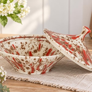 Puede incluir: Un tajine de cerámica con una base de color crema y un patrón moteado de salpicaduras de pintura roja vibrante y marrón oscuro. La tapa cónica está separada y descansa junto al cuenco, mostrando el mismo diseño de salpicaduras rojas y marrones.