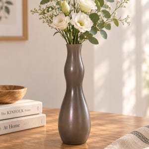Peut inclure: Un vase haut et élancé de couleur bronze métallique avec une silhouette incurvée en forme de sablier. Le vase contient un bouquet de fleurs d'eustoma blanches, de petites fleurs blanches de remplissage et des feuilles d'eucalyptus vertes.
