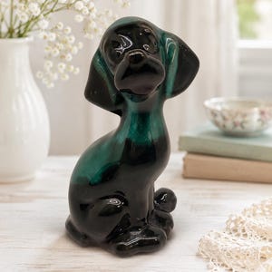Può includere: Una statuetta di cane in ceramica con finitura lucida e una sfumatura di colore che passa dal nero profondo alla base a un vivace verde smeraldo sulla parte superiore del corpo e sulla testa, raffigurata in posizione seduta con lunghe orecchie.