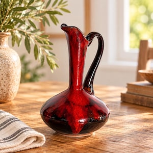 Peut inclure: Un pichet en céramique brillante avec un vernis marbré rouge et noir éclatant. L'objet présente une base large et arrondie, un col élancé et une anse incurvée partant du bord vers l'épaule, avec un bec verseur asymétrique et évasé.