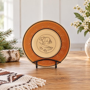 Peut inclure: Une assiette ronde en céramique avec un centre crème orné d'un dessin au trait noir représentant un ours, un poisson et des empreintes de pattes. Le bord est d'une teinte orange-brun chaude avec un motif répétitif de petits bonshommes noirs.