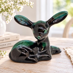 Könnte beinhalten: Eine Keramikfigur eines ruhenden Hirsches mit großen, aufrechten Ohren. Die Skulptur ist mit einer glänzenden, tiefschwarzen Glasur überzogen, die an den Ohren und am Rücken lebendige smaragdgrüne Akzente aufweist und eine glatte Oberfläche bildet.