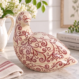 Può includere: Una statuetta di uccello in ceramica color crema caratterizzata da un intricato motivo a volute rosso scuro su tutta la superficie. L'uccello è raffigurato in posizione seduta con la testa leggermente girata di lato e finitura liscia.