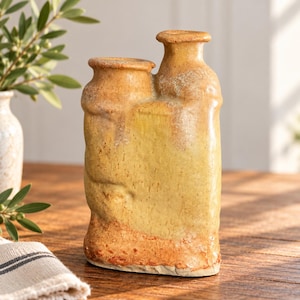 Könnte beinhalten: Eine doppelhalsige Keramikvase mit einer texturierten, gesprenkelten gelben und orangen Glasur. Das Gefäß verfügt über zwei unterschiedliche Öffnungen in verschiedenen Höhen und eine raue, unebene Oberfläche mit Farbvariationen.